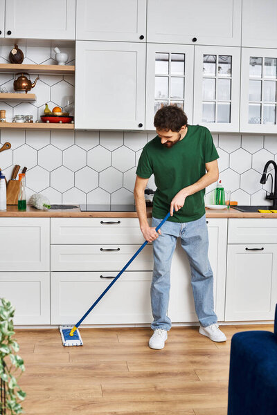 handsome joyous man in comfortable casual attire using mop to clean floor while in kitchen at home