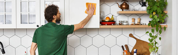 back view of adult man in dark green t shirt using rag to clean kitchen cupboards at home, banner