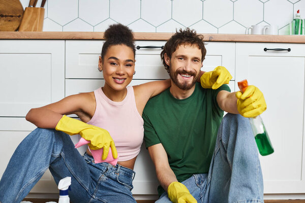 joyous attractive diverse couple in cozy casual outfits sitting on floor and smiling at camera