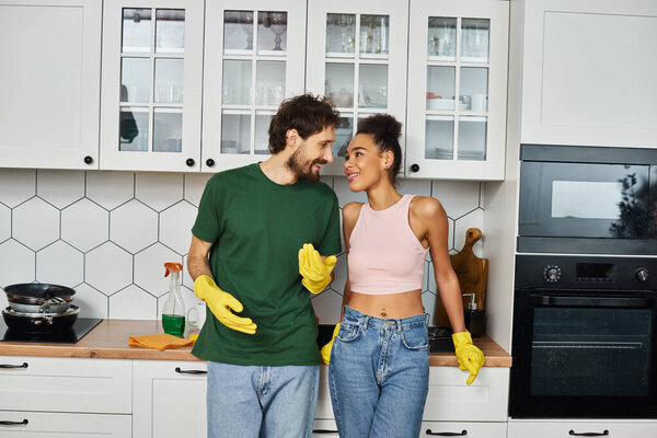 appealing multicultural jolly couple in casual attires smiling at each other during spring cleaning