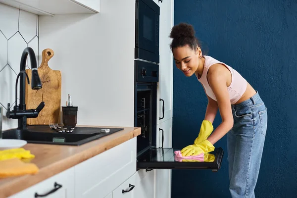 快乐的非洲裔美国女人在家里穿衣 在家里用抹布洗她的现代烤箱 在春天打扫卫生 — 图库照片