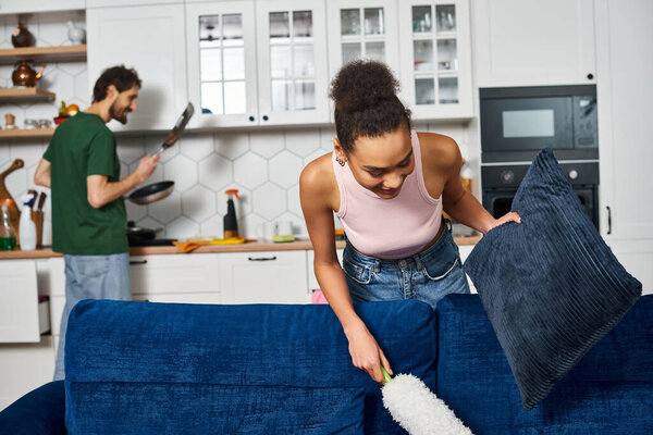 focus on joyous african american woman using duster with her blurred jolly boyfriend on backdrop
