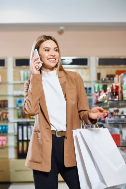 Güzel görünümlü, neşeli, modaya uygun giyinmiş, kozmetik mağazasında telefonla konuşan bir kadın.