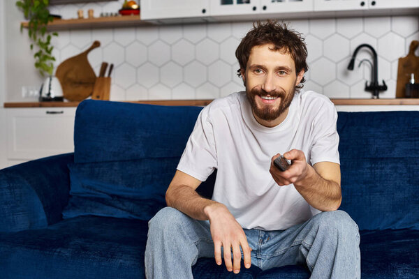 handsome joyous man in comfy homewear watching TV and holding remote control while at home