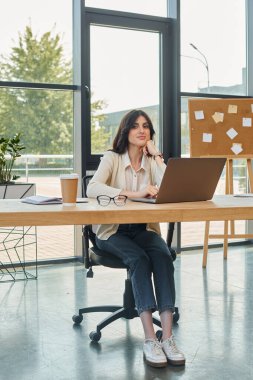 Modern bir iş kadını masasında oturur, modern ofis ortamında dizüstü bilgisayarına odaklanır..