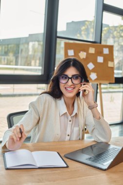 Profesyonel bir iş kadını, bayilik kavramının bir parçası, modern bir ofiste masa başında oturuyor, cep telefonuyla konuşuyor..