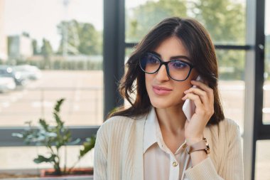 Bir iş kadını gözlük takıyor, modern ofis ortamında cep telefonuyla konuşuyor, bayilik konseptini sergiliyor..