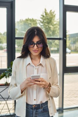 Odaklanmış bir iş kadını pencerenin kenarında duruyor, modern bir ofis ortamında cep telefonunu kontrol ediyor..