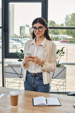 Odaklanmış bir iş kadını modern bir ofiste masada duruyor, telefonuna dalmış, bayilik kavramlarını düşünüyor..