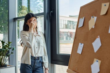 Modern bir ofiste yorgun bir iş kadını pencerenin önünde duruyor.,