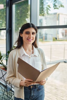 Bir iş kadını, elinde bir klasör, bir bayilik ofisindeki modern bir CEO 'nun özünü somutlaştırarak, bir pencerenin önünde güvenle durur..