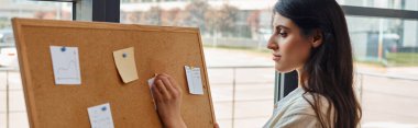 Bir iş kadını, modern ofis çalışma alanındaki renkli not kağıtlarıyla süslenmiş bir ilan tahtasını araştırıyor..