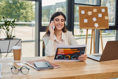 Bir iş kadını, modern bir ofis ortamında telefon konuşmasına dalmış bir şekilde masada oturuyor..