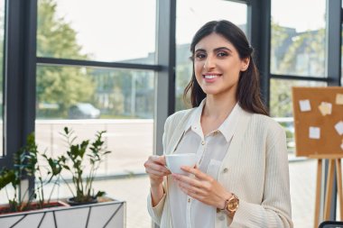 A businesswoman savors a cup of coffee against a cityscape backdrop through a large window in a modern office. clipart