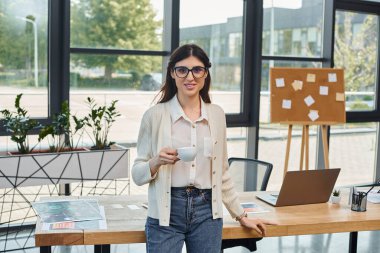 A businesswoman stands in a modern office, focused on her laptop on the table in front of her. clipart