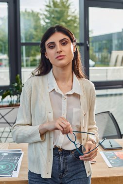 Modern bir iş kadını, bir şirket ofisinde, parlak bir masanın önünde elinde bir çift gözlük tutarak duruyor..