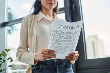 Bir iş kadını pencerenin kenarında duruyor, modern ofis ortamında bir kontrat tutuyor..