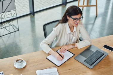 A businesswoman sits at a table in a modern office, focused on her laptop, working on a franchise concept. clipart