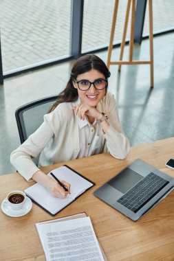 A businesswoman is deep in thought, sitting at a table with a notebook and pen in a modern office setting. clipart