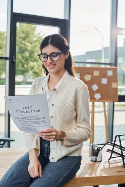 A businesswoman sitting on a table, confidently holding a piece of paper in a modern office setting near her workspace. clipart