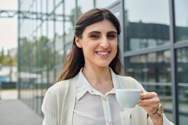 Modern bir binanın önünde elinde bir fincan kahve tutan şık bir iş kadını..