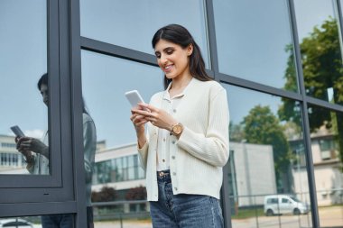 Odaklanmış bir iş kadını bir binanın önünde durup, düşünceli bir ifadeyle telefonunu karıştırıyor..