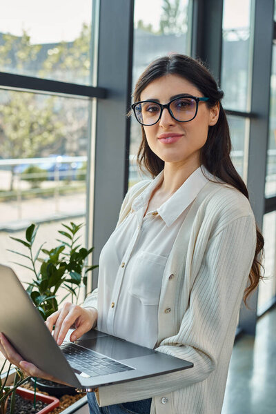 A sophisticated woman in glasses confidently holds a laptop in a modern office, embodying innovation in the business world.