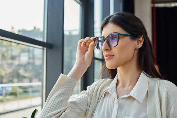 A businesswoman, wearing glasses, gazes out a window in a modern office, contemplating the urban landscape.