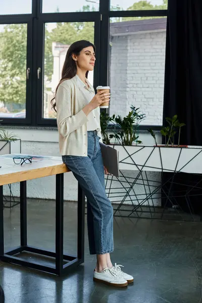 stock image A modern businesswoman stands at a table, enjoying a cup of coffee in a sleek office setting.