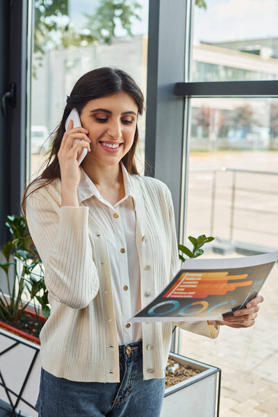 Деловая женщина многозадачная, разговаривая по мобильному телефону, держа карты в современной офисной обстановке.