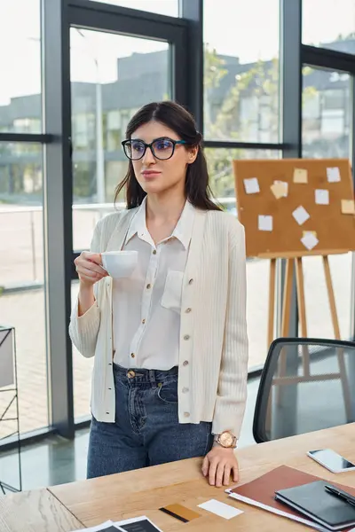 ビジネスウーマンは フランチャイズコンセプトに焦点を当てた机の前にカップを持って現代のオフィスに立っています — ストック写真