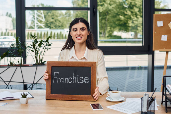 A modern businesswoman sits confidently at a table, holding a sign with a purposeful expression.