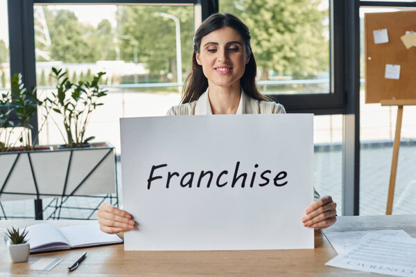 A businesswoman in a modern office sitting at a table and holding a sign with franchise letters