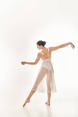 Genç bir kadın uzun beyaz bir elbiseyle zarif bir şekilde dans ediyor. Beyaz arka planda bir stüdyoda güzel ve zarif bir şekilde dans ediyor..