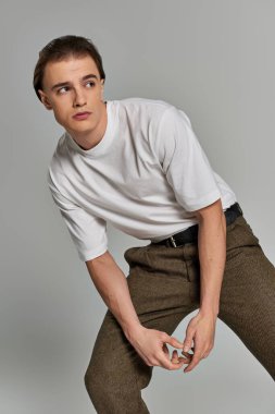 Tişörtlü ve kahverengi pantolonlu yakışıklı genç adam gri arka planda çekici bir şekilde poz veriyor ve başka tarafa bakıyor.