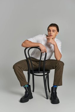 Moda kıyafetleri içinde çekici bir genç sandalyede oturuyor ve gri arka plana bakıyor.