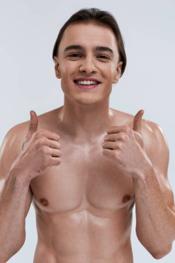 Neşeli, seksi, üstsüz, başparmakları havada poz veren ve kameraya mutlu bir şekilde gülümseyen adam.