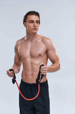 Çekici, üstsüz, siyah pantolonlu genç adam fitness genişletici ile antrenman yapıyor ve başka tarafa bakıyor.