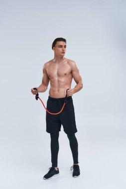 Çekici, üstsüz, siyah pantolonlu genç adam fitness genişletici ile antrenman yapıyor ve başka tarafa bakıyor.