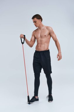 alluring shirtless sporty young man in black pants training with fitness expander and looking away clipart