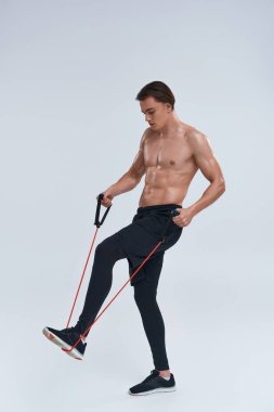 Çekici, üstsüz, siyah pantolonlu genç adam fitness genişletici ile antrenman yapıyor ve başka tarafa bakıyor.