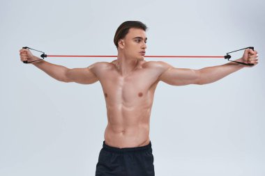 Çekici, üstsüz, siyah pantolonlu genç adam fitness genişletici ile antrenman yapıyor ve başka tarafa bakıyor.