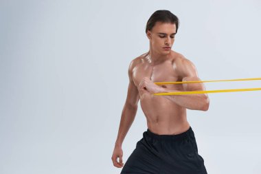 Siyah pantolonlu genç sporcu, gri arka planda direniş bandıyla egzersiz yapıyor.