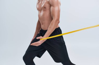 Siyah pantolonlu atletik gömleksiz seksi adamın gri arka planda direnç bandını kullanması.
