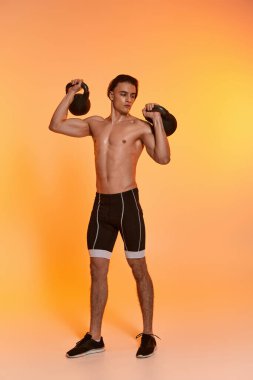 handsome sporty shirtless man exercising with kettlebells and looking away on orange backdrop clipart