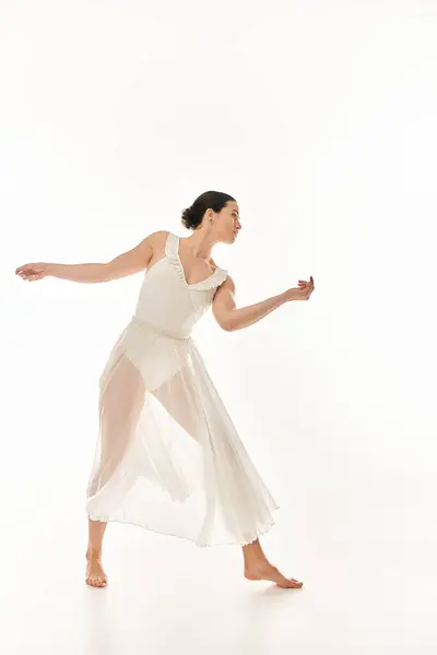 若い女性は 白い背景に対して設定されたスタジオで流れる白いドレスで踊るように優雅さと優雅さを誇ります — ストック写真