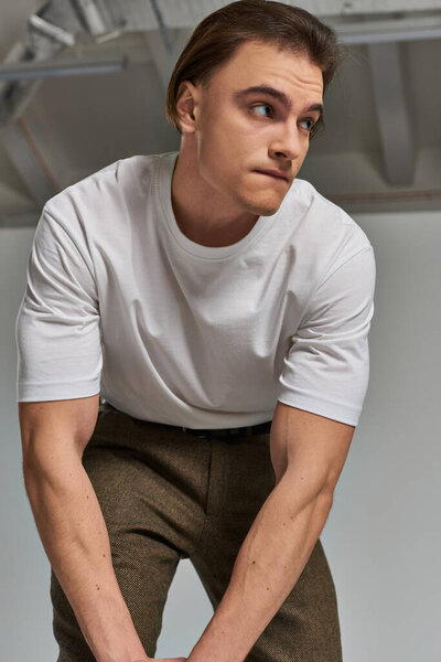 appealing young man in t shirt and brown pants posing attractively on gray backdrop and looking away