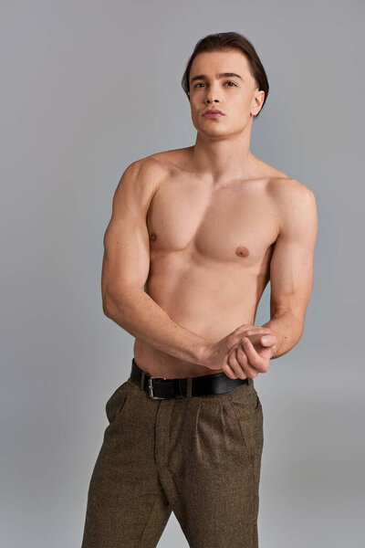 alluring shirtless man in brown elegant pants posing attractively on gray backdrop and looking away