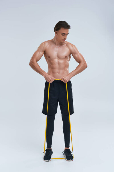 alluring athletic young man in black pants posing topless with resistance band and looking away