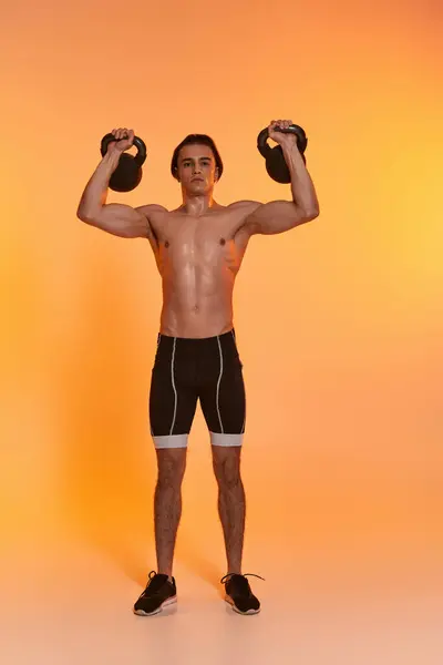 stock image appealing sporty shirtless man exercising with kettlebells and looking at camera on vivid backdrop